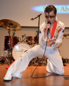 DELVIS (teen singer and Elvis Entity) wearing dark sunglasses gets down on one knee as he sings an Elvis Presley song.